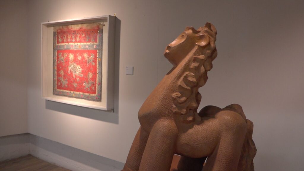 Escultura de animal estilizado en primer plano dentro de una sala de museo, con un cuadro rojo enmarcado colgado en la pared al fondo.