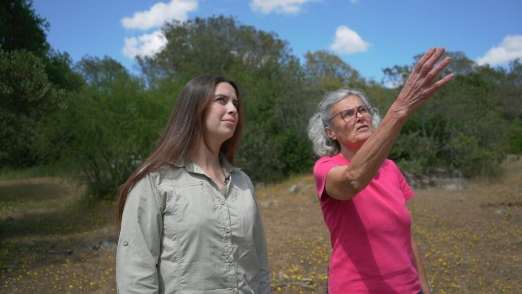 Dos mujeres de pie en un entorno natural: una mujer mayor señala hacia el horizonte mientras explica algo, y una mujer más joven observa atentamente; al fondo hay vegetación, suelo seco y cielo despejado.