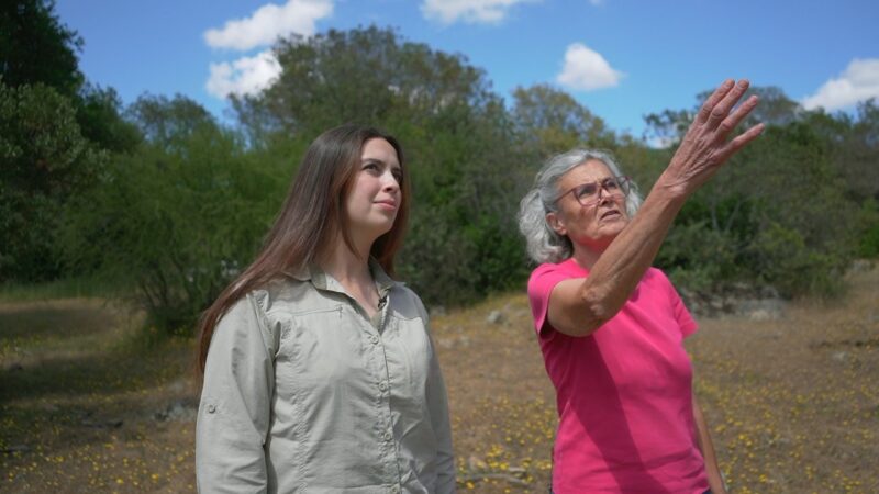 Dos mujeres de pie en un entorno natural: una mujer mayor señala hacia el horizonte mientras explica algo, y una mujer más joven observa atentamente; al fondo hay vegetación, suelo seco y cielo despejado.