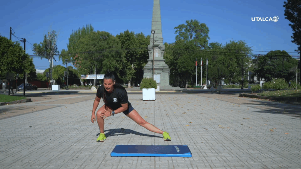 Persona realizando ejercicios de fuerza sobre una colchoneta azul en una plaza al aire libre, rodeada de árboles y áreas verdes.