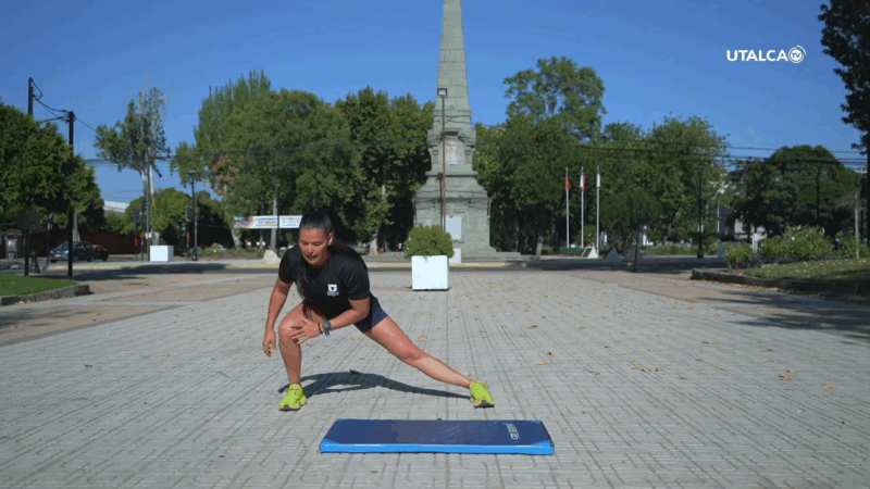 Persona realizando ejercicios de fuerza sobre una colchoneta azul en una plaza al aire libre, rodeada de árboles y áreas verdes.
