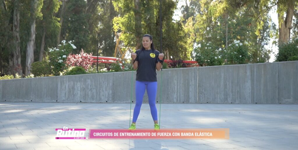 Persona realizando ejercicios de fuerza con banda elástica al aire libre, ejecutando una flexión de brazos de pie para fortalecer bíceps y tren superior.