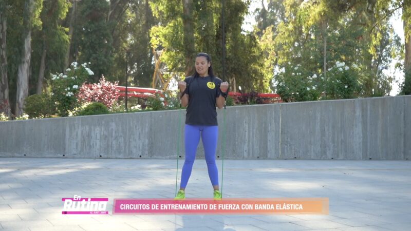 Persona realizando ejercicios de fuerza con banda elástica al aire libre, ejecutando una flexión de brazos de pie para fortalecer bíceps y tren superior.
