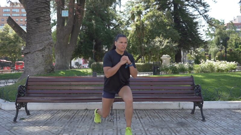 Persona realizando una zancada asistida frente a una banca en una plaza, como parte de una rutina de ejercicio al aire libre.